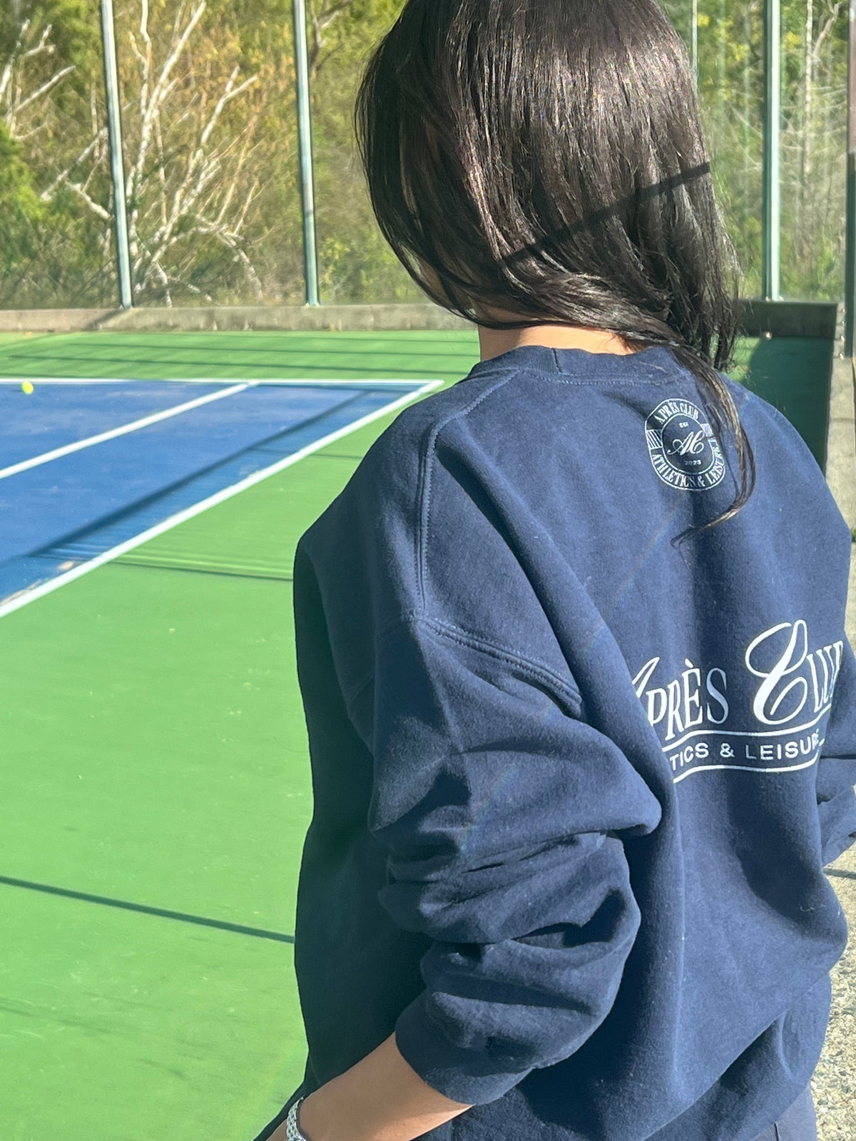 Navy Après Club crest sweatshirt worn on a tennis court, The Clubhouse collection
